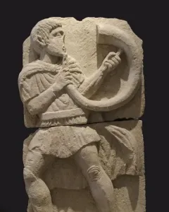 Broken stone relief of the Acrobat from Osuna showing a male figure playing a horn.