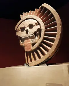 Disk of Mictlāntēcutli stone sculpture with a skull-like face and semicircular ribbed frame in a museum display.