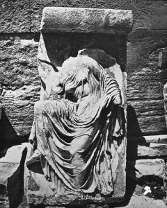 Damaged relief sculpture of Nike Untying Her Sandal, shown headless and draped in flowing robes beside a stone wall.