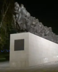 The Train of Pain monument at night, showing a long dark sculpture of clustered human figures on a light-colored base.