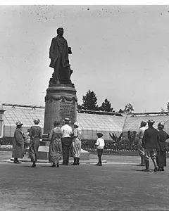 William Henry Seward statue on a tiered pedestal in Volunteer Park, with people gathered nearby and glass greenhouses in the background.