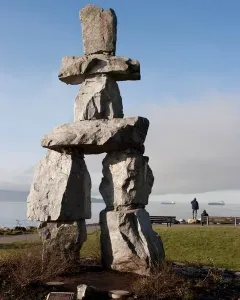 Inukshuk stone monument beside a grassy shoreline with water and distant ships under a clear blue sky.