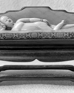 Doctor\'s Lady, a light-colored carved female figure reclining on a patterned daybed with an upholstered back.