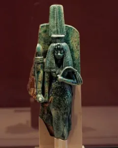 Greenish-blue statue of Queen Tiye wearing a tall ornate headdress and long textured garment, holding objects in both hands.