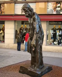 Eustache de Saint Pierre bronze sculpture of a bearded man in long robes standing on a stone base beside storefront windows