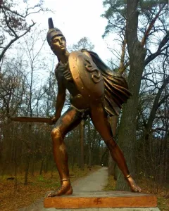 Bronze Greek warrior monument showing a helmeted soldier with a shield and raised sword.