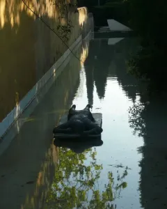 Ophelia Drowned reclining on a platform in a shallow canal beside greenery and a sunlit wall.