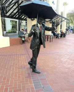 Allow Me, a dark bronze sculpture of a man in a suit holding a large umbrella and raising one hand.