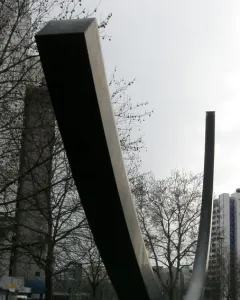 Bernar Venet’s Arc de 124,5° sculpture rising over a grassy area with bare trees and city buildings in the background.