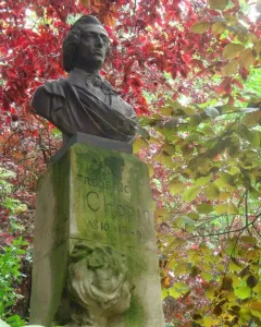 Dark metallic bust of Frédéric Chopin mounted on a weathered stone block amid red and green foliage.