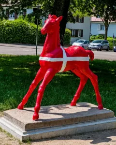 Holbeinpferd sculpture painted bright red with a white cross, standing on a concrete base in a grassy area.