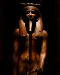 Wooden statue of King Hor with a pharaonic headdress and false beard on a black background.