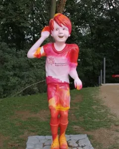 Schängel statue of a smiling boy with one hand raised to his ear, painted in red, pink, and yellow patterns.