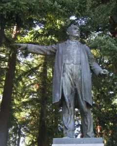 Harvey W. Scott statue of a man in a long coat pointing forward, set on a light-colored base among pine trees.