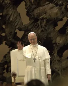 Pope Leo XIV in white clerical vestments stands in front of The Resurrection sculpture, smiling and waving.