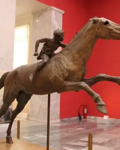 Jockey of Artemision bronze sculpture showing a young rider leaning forward on a horse in motion inside a museum.