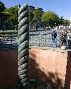 The Serpent Column with a weathered green patina stands inside a shallow red-walled enclosure near an ornate fence and public square.