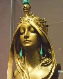 Polished bronze bust of a woman with closed eyes, wearing an ornate crown and long teal earrings.