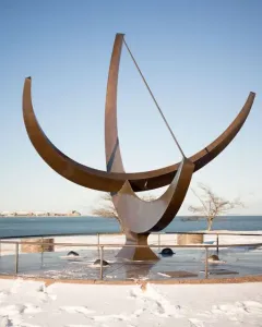 Man Enters the Cosmos sundial sculpture on a circular base beside a snowy lakeshore, with a city skyline in the distance.