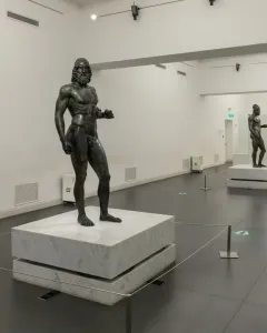 The Riace bronzes displayed on white pedestals in a brightly lit museum hall.