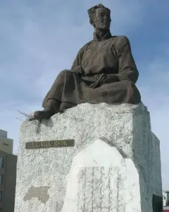 Natsagdorj Monument showing a weathered bronze-colored seated figure on a stone pedestal with engraved inscriptions and a floral wreath at the base.