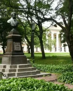 Bust of Pushkin on a tall stone pedestal in a park, with trees and a columned building in the background.