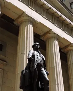 The Statue of George Washington stands on a stone platform before a classical building with tall fluted columns.