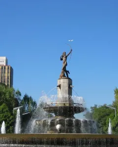 Diana the Huntress Fountain with a figure drawing a bow against a clear sky and city skyscrapers.