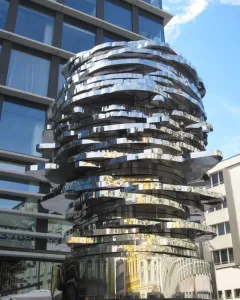 The Head of Franz Kafka sculpture with stacked, mirrored metal panels reflecting nearby buildings and blue sky.