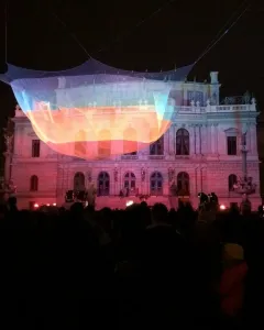 1.26 illuminated mesh sculpture glowing in bands of orange, yellow, and blue above a crowd near the Rudolfinum at night.