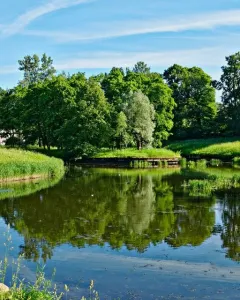 A slender monument to Elena Pavlovna stands near a multi-arched stone bridge over calm water surrounded by green trees.