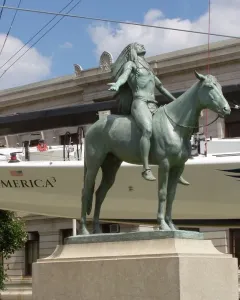 Appeal to the Great Spirit bronze statue of a Native American rider on horseback with arms outstretched, set against a sailboat and stone building.
