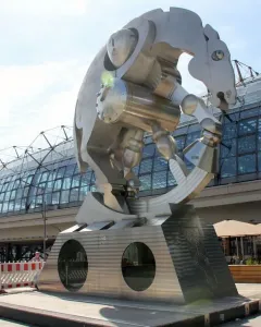 Rolling Horse sculpture beside a modern station with a vaulted glass roof, shown on a wedge-shaped pedestal with round porthole windows.