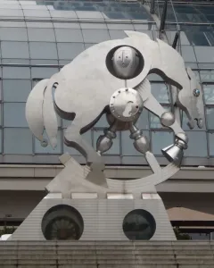 Rolling Horse sculpture beside a modern station with a vaulted glass roof, shown on a wedge-shaped pedestal with round porthole windows.
