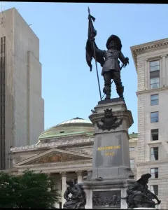 Maisonneuve Monument with a bronze figure holding a flag and sword atop a stone pedestal inscribed “MAISONNEUVE 1642,” with smaller figures below and city buildings in the background.