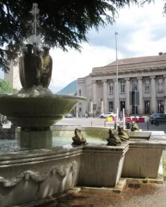 The Fountain of the Frogs stone fountain sprays water in a city square with a large classical building in the background.