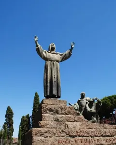 Bronze Monument to Saint Francis of Assisi with robed figures on a stepped stone plinth under a blue sky.