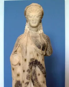 Chiotissa kore sculpture showing a female figure with braided hair and patterned draped garments on a red pedestal.