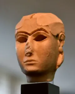 The Warka Mask, a light-toned ancient stone head with stylized facial features and hollow eye sockets on a dark pedestal.