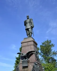 Monument of Alexander III on an ornate pedestal with a decorative iron fence in front of a classical building.