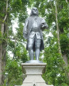 Benjamin Franklin statue in colonial attire on an ornate stone pedestal, framed by trees.