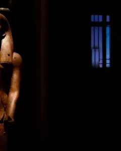 Wooden statue of King Hor with a pharaonic headdress and false beard on a black background.
