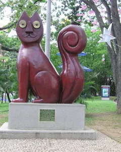 El Gato del Río cat sculpture on a stone pedestal in a park, with a small bird perched on its curled tail.