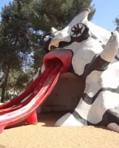 The Golem sculpture with a red mouth and three red slide chutes on sandy ground under a blue sky.