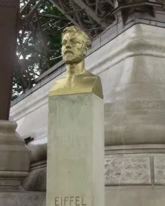 Golden bust of Gustave Eiffel on a stone pedestal with the Eiffel Tower framework in the background.