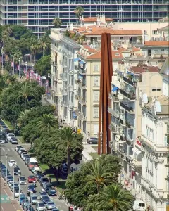 Neuf Lignes Obliques sculpture standing between apartment buildings on a busy city street lined with parked cars and palm trees.