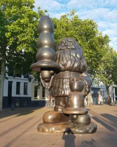 Bronze Santa Claus statue in a plaza, holding an abstract object in one hand and a bell-shaped object in the other.