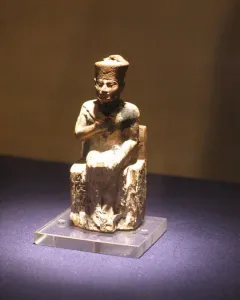 Khufu statuette of a seated figure wearing a tall crown on a clear base.
