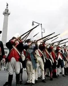 The Trenton Battle Monument rises above colonial reenactors firing muskets in the foreground.