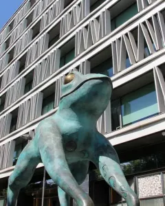 Lucky Frog statue with teal patina and golden eyes in front of a modern building.